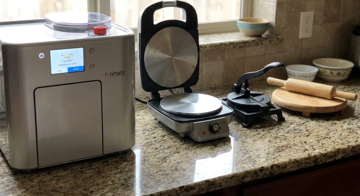 A Rotimatic robot, an electric roti press and a hinged cast-iron press lined up on a kitchen counter beside a wooden board and rolling pin