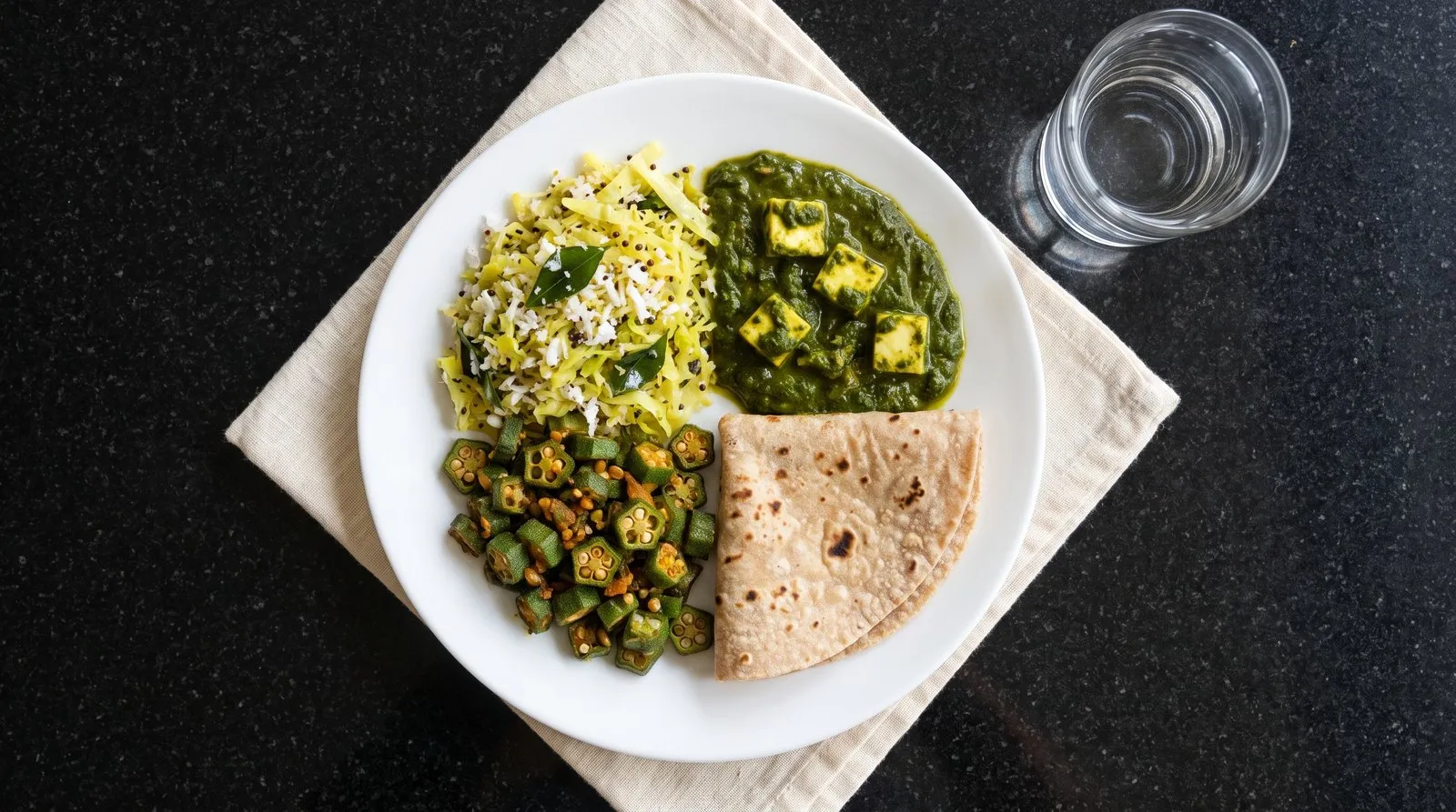 A white plate built the plate-method way: half cabbage thoran and okra, a quarter palak paneer and a quarter one folded whole-wheat roti, with a glass of water