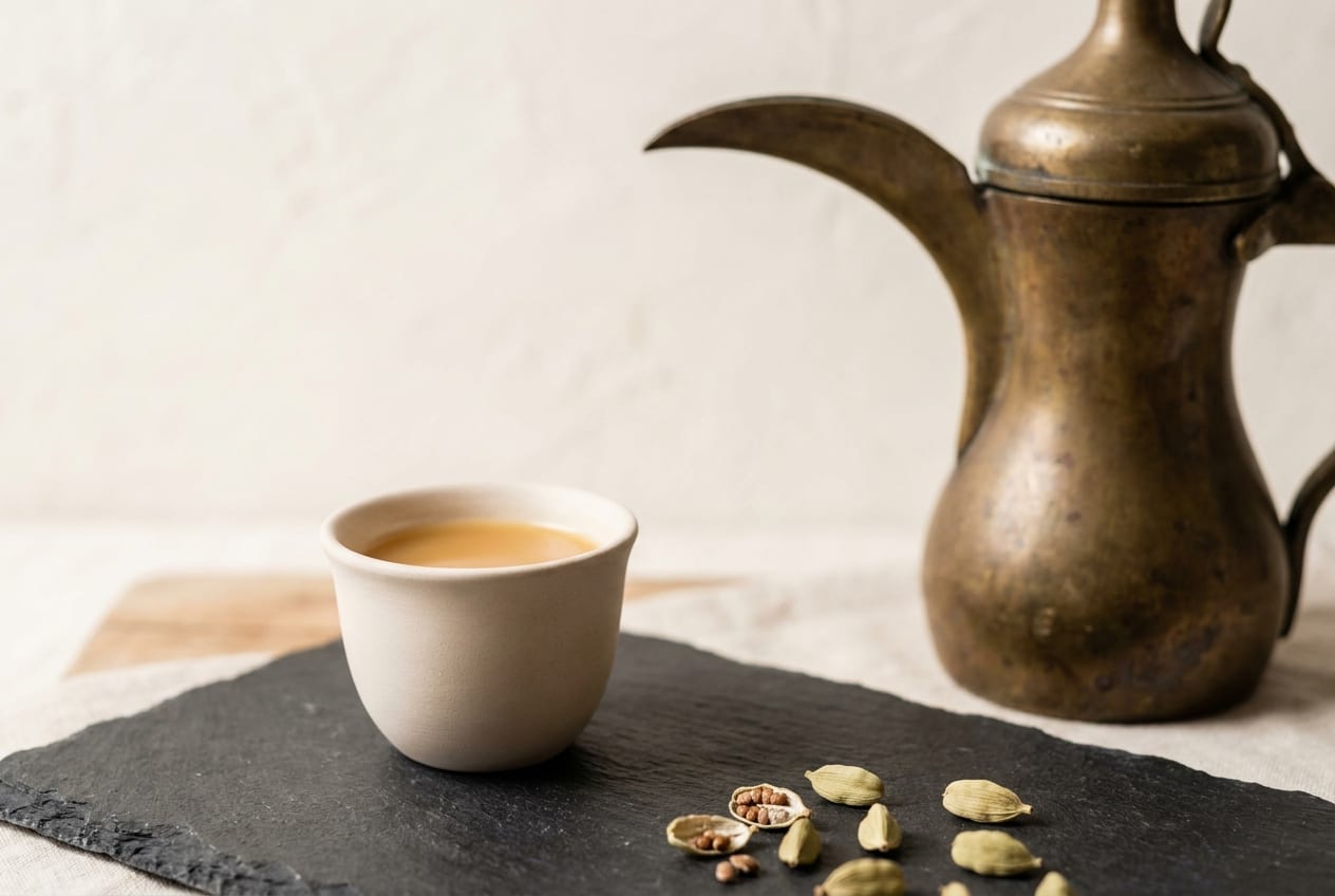 A small handleless cup of pale cardamom coffee beside a brass dallah pot and split green cardamom pods on dark slate