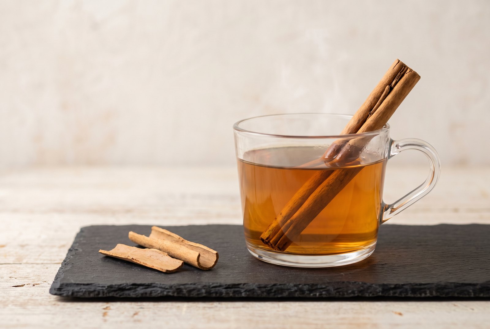 A clear glass cup of amber cinnamon tea with a pale Ceylon cinnamon quill resting in it, on dark slate beside loose cinnamon bark