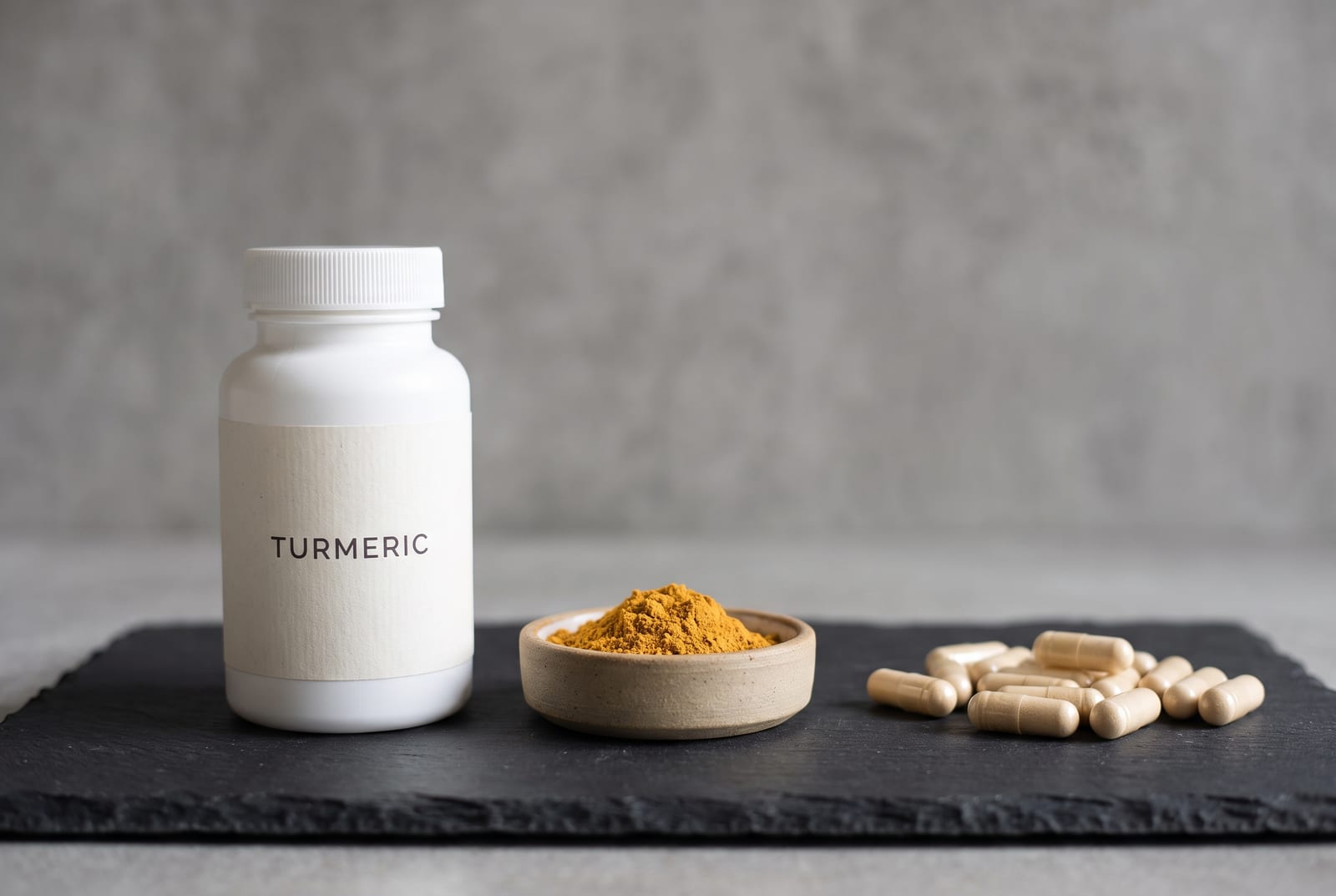 Ground turmeric in a small dish beside scattered capsules on a dark slate surface